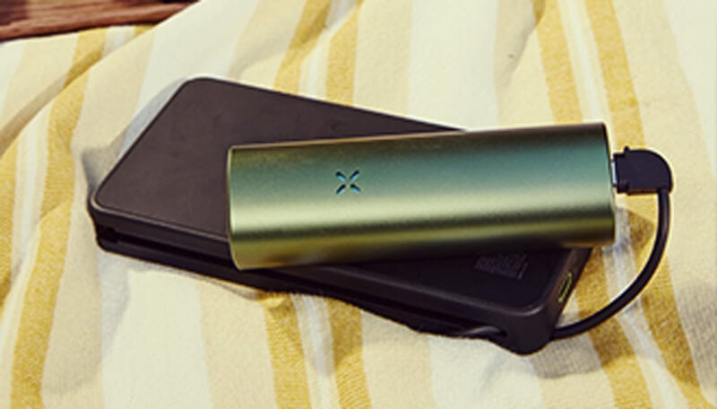 Opladen van de Pax Flow Vaporizer Een slanke groene Pax Flow vaporizer aangesloten op een zwarte powerbank via een USB-kabel, rustend op een geel-wit gestreepte handdoek voor handig opladen onderweg.