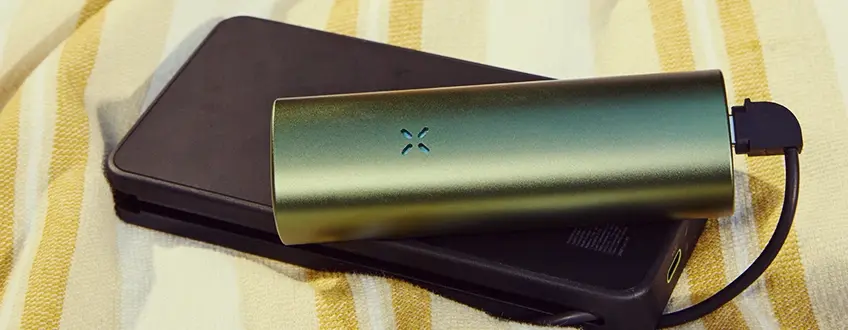 Opladen van de Pax Flow Vaporizer Een slanke groene Pax Flow vaporizer aangesloten op een zwarte powerbank via een USB-kabel, rustend op een geel-wit gestreepte handdoek voor handig opladen onderweg.