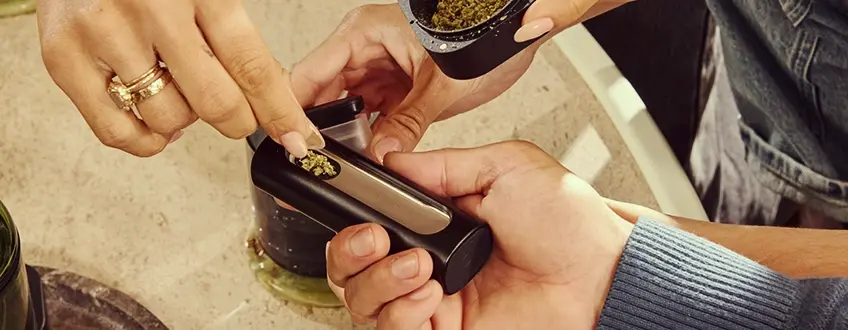 Bijvullen van de Pax Flow Vaporizer Een hand die de kamer van de Pax Flow vaporizer bijvult met vers gemalen cannabis met een klein lepeltje uit een grinder, geplaatst op een marmeren tafel voor nauwkeurig herladen.