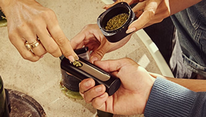 Bijvullen van de Pax Flow Vaporizer Een hand die de kamer van de Pax Flow vaporizer bijvult met vers gemalen cannabis met een klein lepeltje uit een grinder, geplaatst op een marmeren tafel voor nauwkeurig herladen.